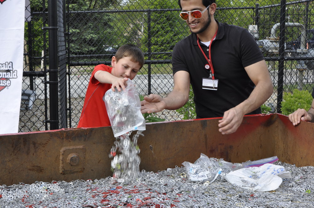 Mohammed standing brhind container of pull tabs, watching child pour tabs from bag.