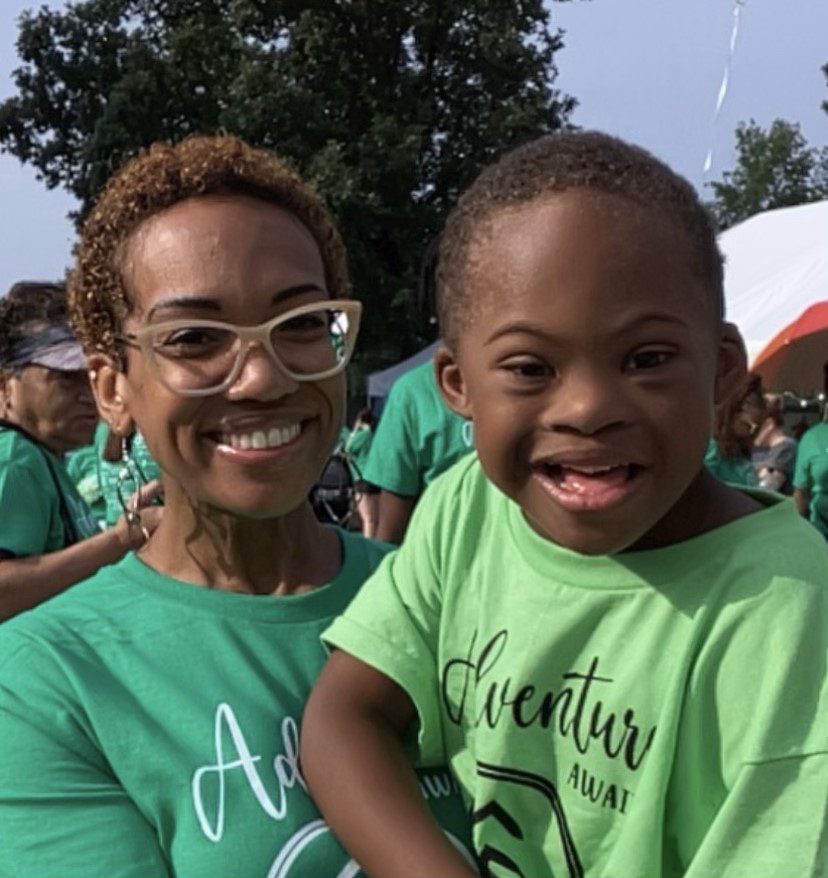 Adelaida and Cameron Peete-Brown. They are at an outside event, Adelaida is holding Cameron up to smile for the camera. He is wearing a green T shirt that says 'adventure awaits'