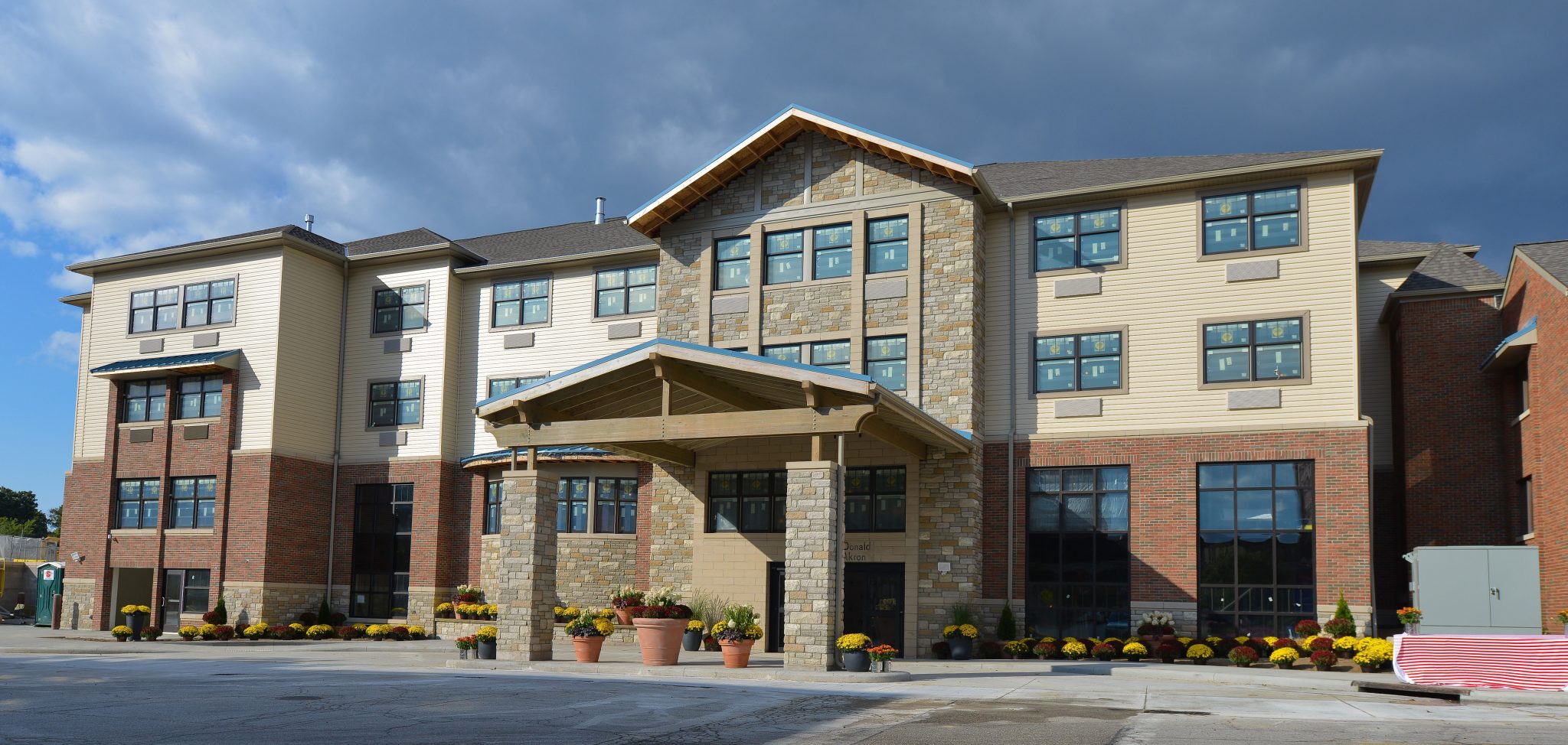 Front exterior of the Akron Ronald McDonald House. It is a four story building with light brown brick on the first 2 floors and covered drive up entrance. The second floor has light yellow siding. There are welcoming flowers and flowerpots along the base of the building.