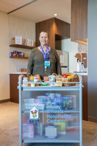 A volunteer smiles behind the Happy Wheels Cart for Family Room at MetroHealth. The cart is loaded up with snacks and drinks for families and their children.