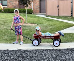 Girls posing for a picture on a sidewalk with a radio flyer red wagon one girl is laying down in the wagon turned toward the camera smiling the other girl is holding the handle And grinning with one hand on her hip