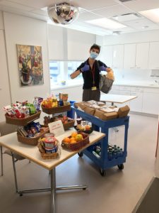 A young man gives a thumbs up behind a cart filed with boxed lunches and water bottles. The table net to him is stocked with baskets of fruit and other snacks.