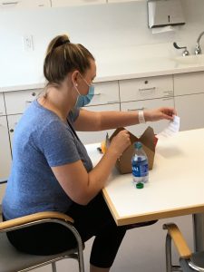 A woman in a blue t-shirt sits at a kitchenette table. She is opening a boxed lunch provided by the RMHC Family Room.