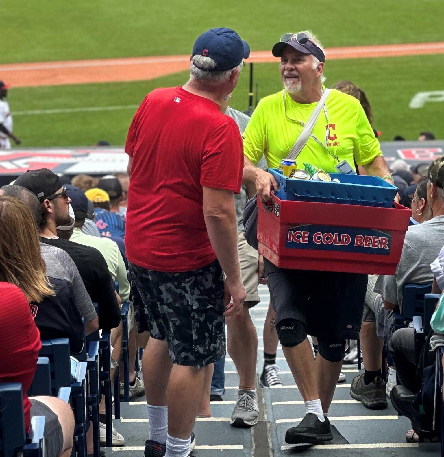 A vendor and a bright yellow shirt carries a tub marked ice cold beer up the stairs at a baseball stadium he has stopped to talk to a man in a red shirt who has his back to the camera