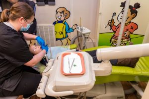 In the Ronald McDonald Care Mobile a child is receiving treatment. The patient reclines in a dental chair colored a cheery bright green. There are cartoons of smiling children on the cream colored walls.