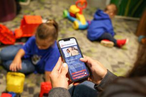 Two children in blue RMHC shirts play on the carpet with toy trucks. In the foreground, a woman's hands are holding a phone displaying the latest version of the RedTreehouse.org website.