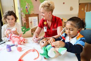 At Akron Children's Beeghly Campus, a STAR Corner Staff member is smiling while she is seated at a table with two young girls playing doctor with baby dolls and toy stethoscopes. 