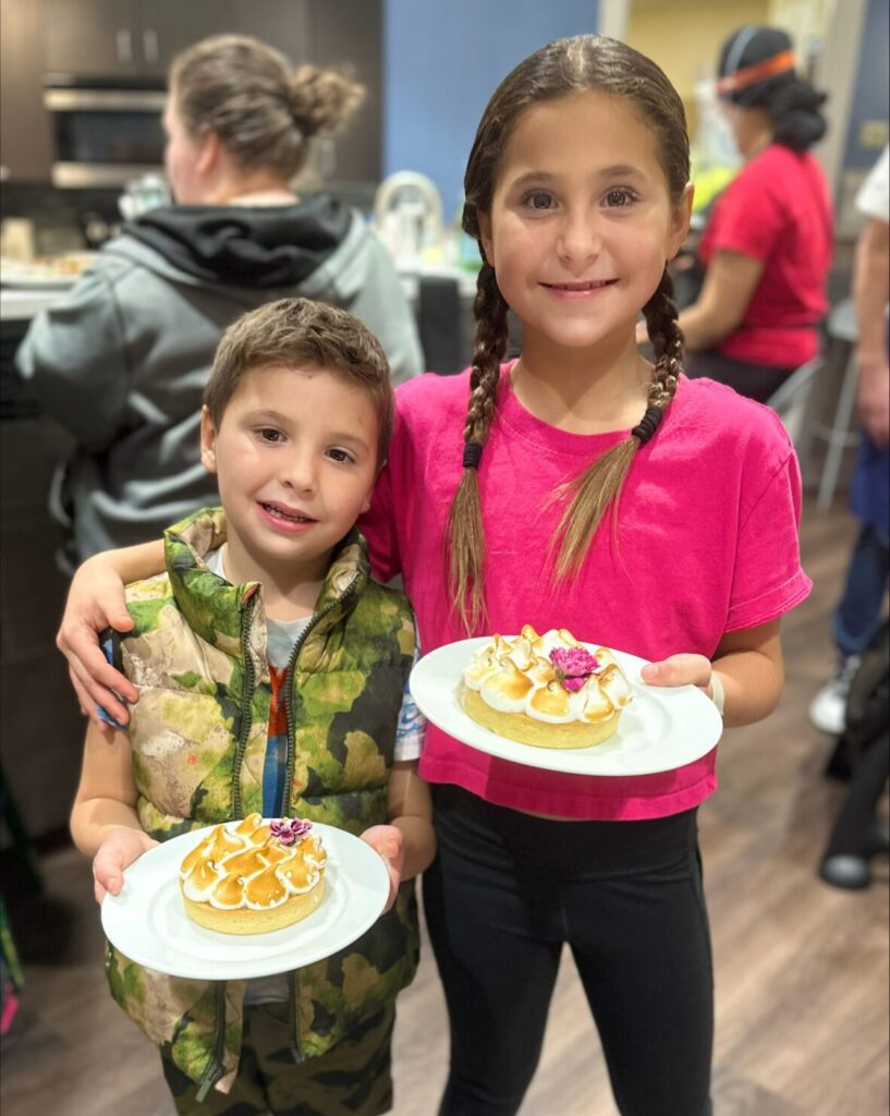 Girl wearing pigtails and pink t shirt smiles with her arm around a young boy wearing a camouflage vest. They are both holding plates with dessert treats.
