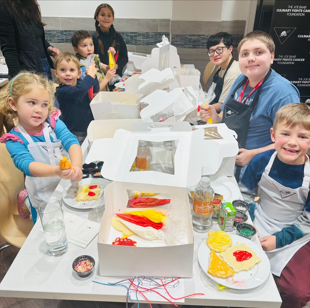 Seven kids are gathered around a long table with white boxes and cookie frosting supplies arranged down the middle