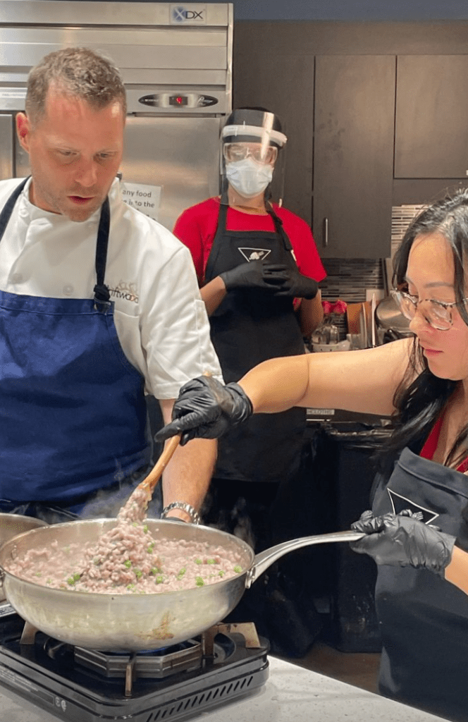 In a commercial kitchen, a young woman is stirring a big wok full of food on a burner. There is a man in a blue apron looking on and another person in a black apron behind them also watching.
