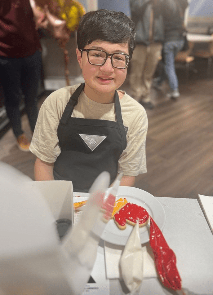 A young person with short dark hair and glasses, wearing an apron and sitting at a table while decorating Christmas cookies