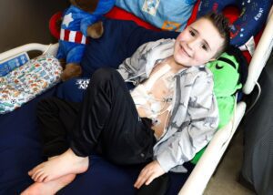 Boy with monitor and bandaged chest scar in hospital bed smiling.