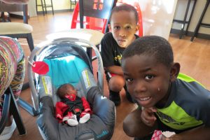 Siblings staying at the Ronald McDonald House. Two young boys crouching next to a tiny infant in a walker. The play area can be seen in the background. 